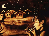 Homokból készült képen János vitéz furulyázik. A háttérben egy falu házai, fölöttük a Hold és a csillagok .